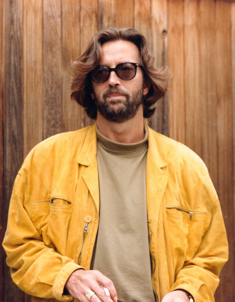 Spevák a gitarista Eric Clapton v žltej bunde s cigaretou v ruke pred drevenou stenou. 